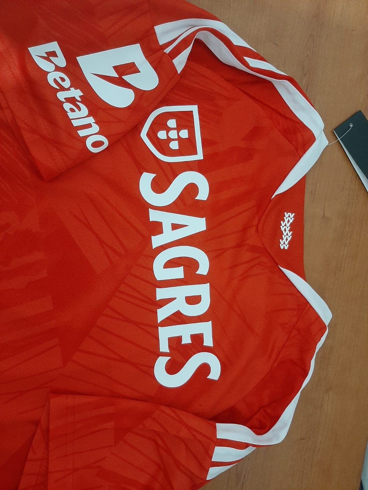 Pormenor do ombro da camisola do SL Benfica 26/27 com as três riscas brancas e os patrocínios Sagres e Betano.